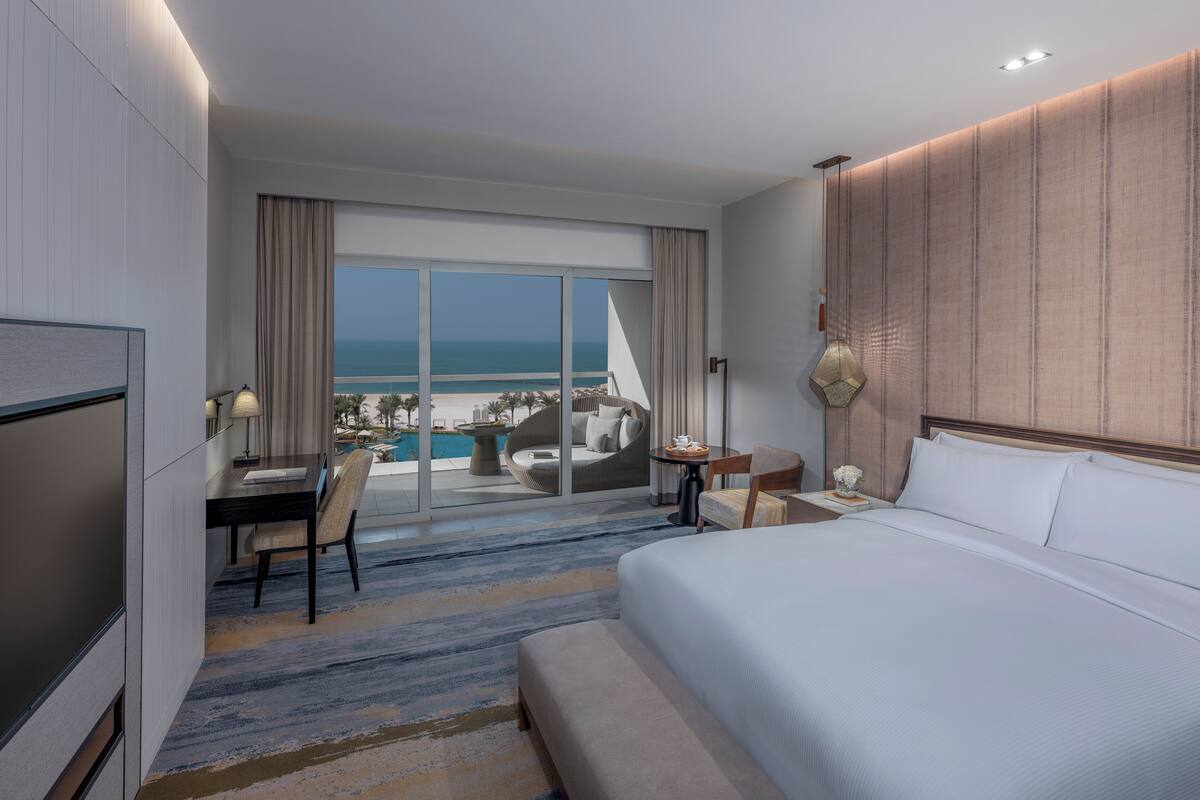 A hotel room with a large bed, a desk with a chair, a small table with a lamp, and a view of the ocean and beach.