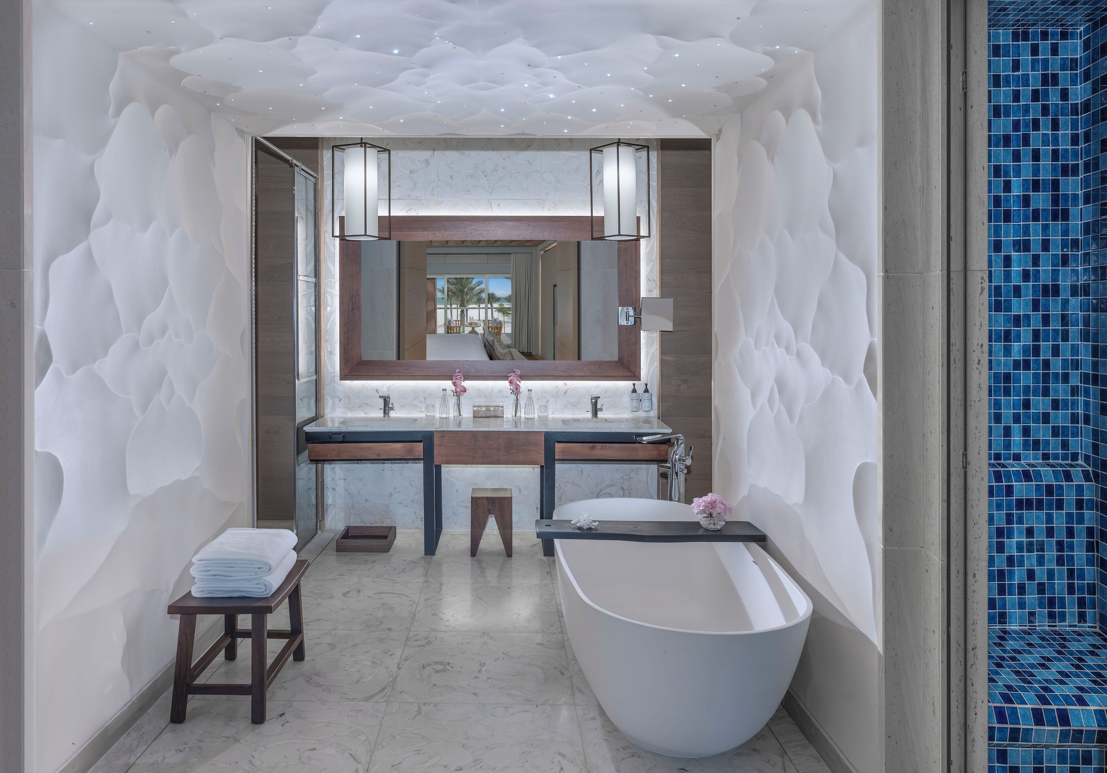 A modern bathroom with a freestanding bathtub, a double vanity with a large mirror, and a small stool with towels.