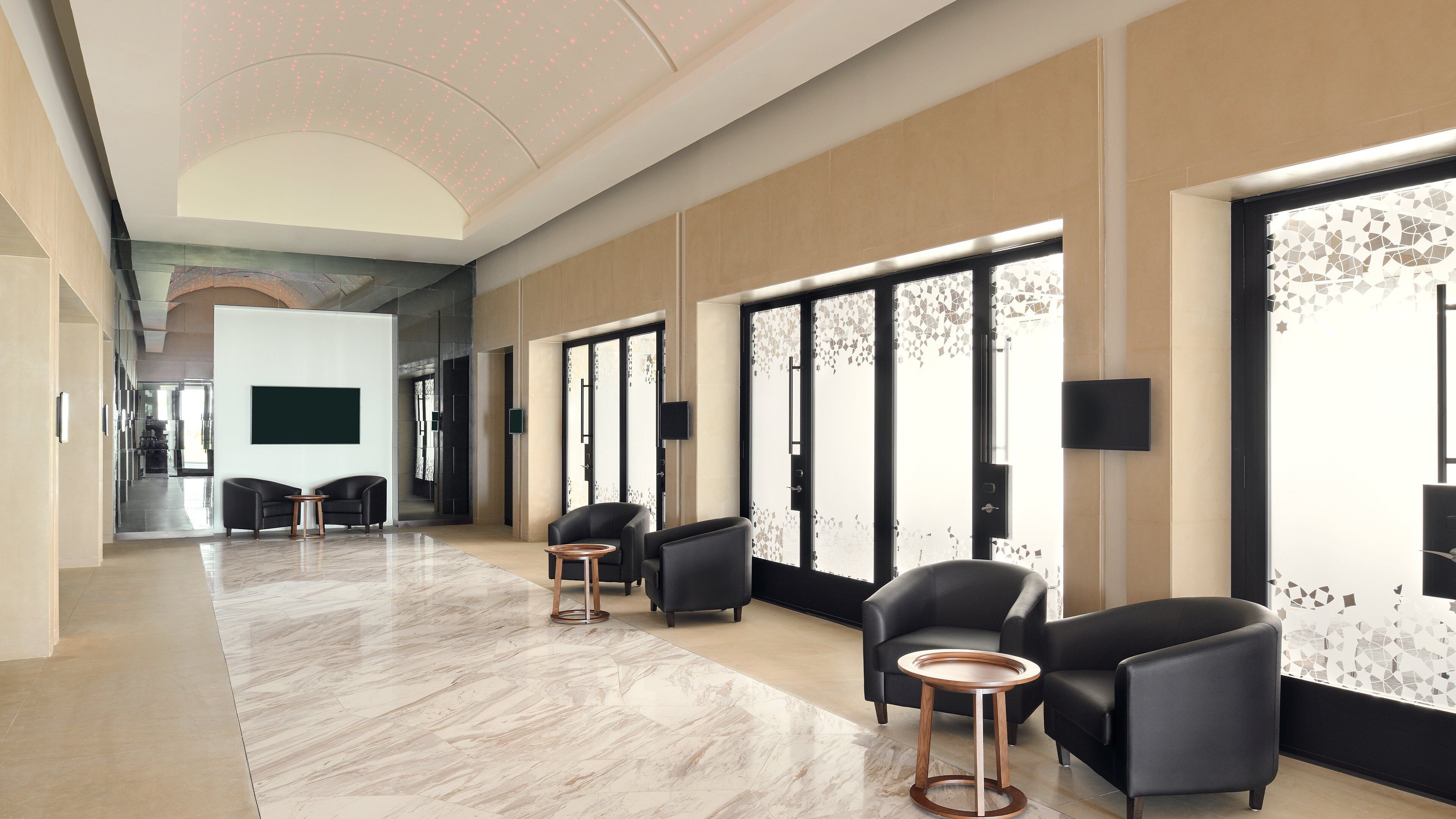 Un hall d’hôtel moderne avec un sol en marbre, des sièges en cuir noir et de grandes fenêtres ornées de panneaux de verre décoratifs.