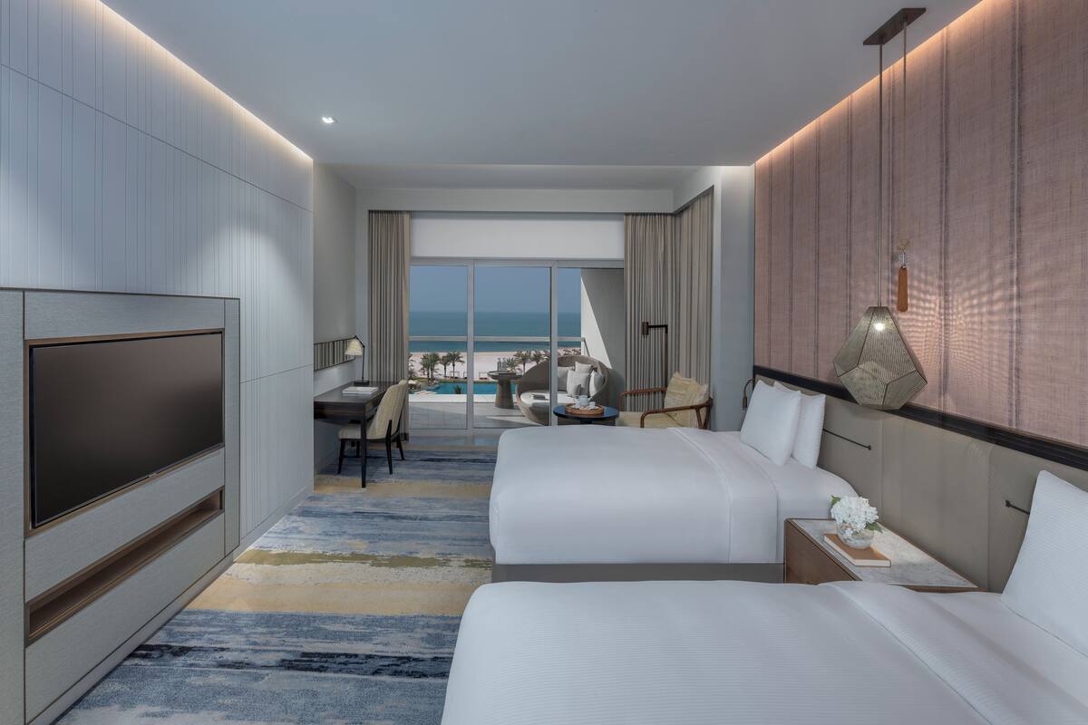 A modern hotel room with a large TV, a desk, two beds, and a view of the beach.
