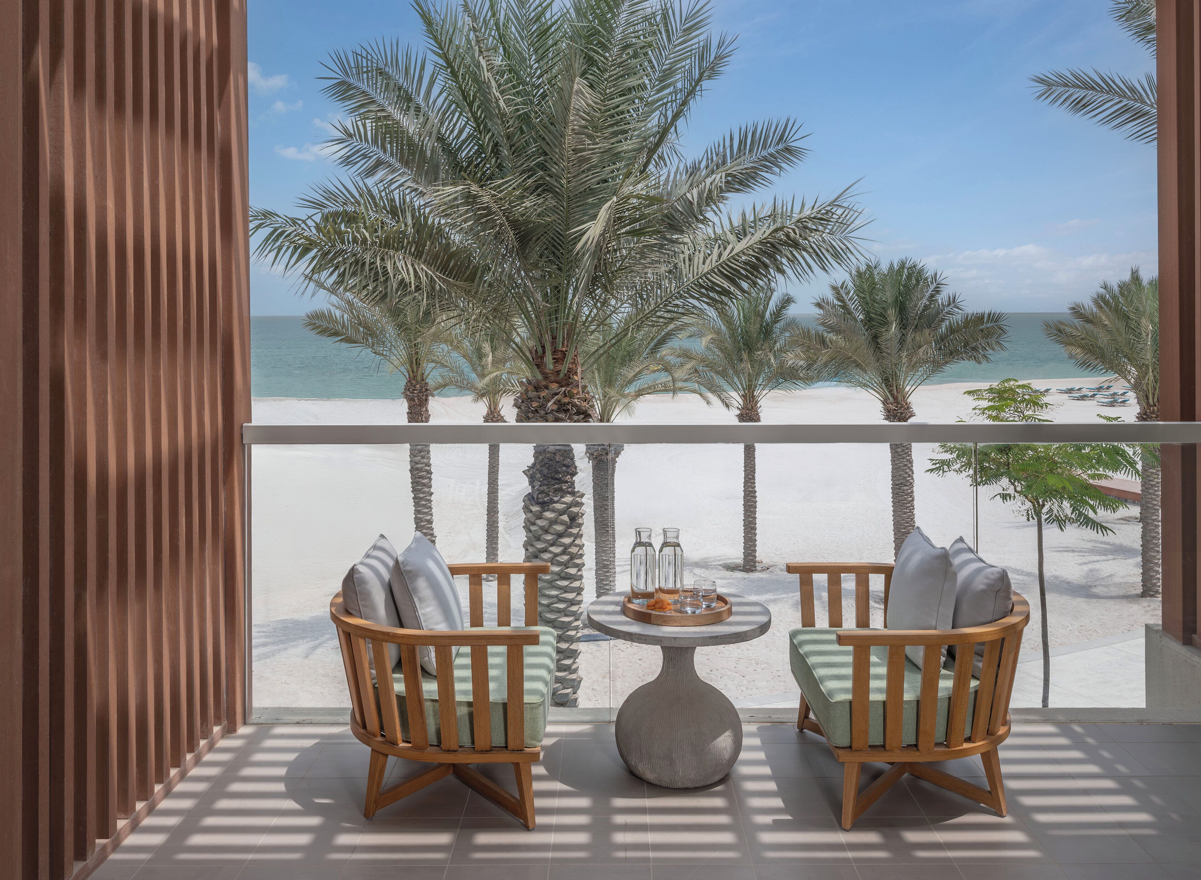 A balcony with two wooden chairs and a round table, overlooking a beach with palm trees and the sea.