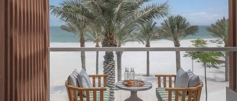 A balcony with two wooden chairs and a round table, overlooking a beach with palm trees and the sea.