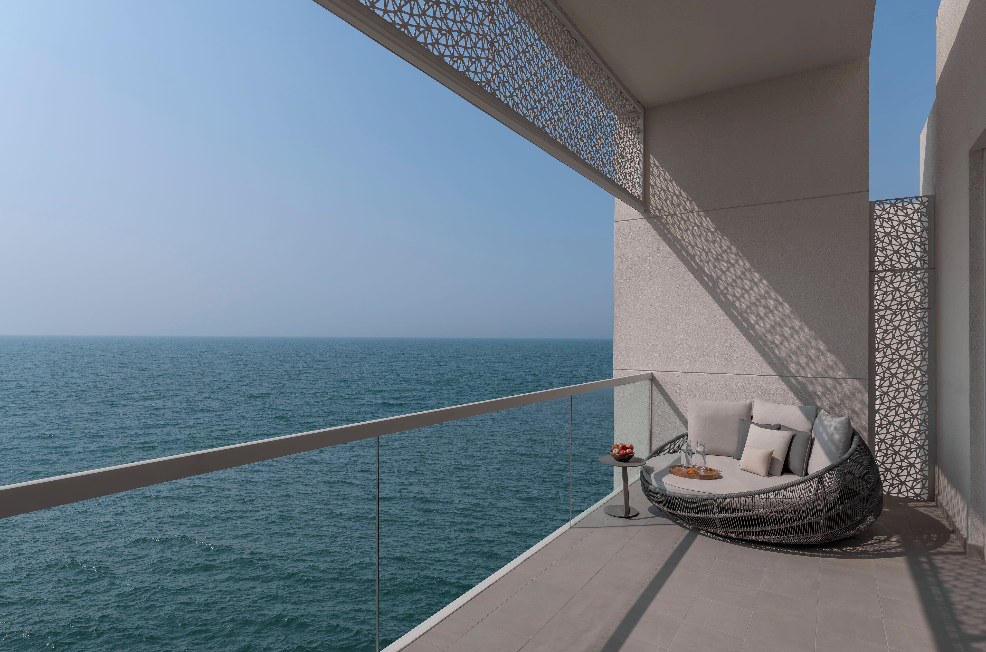 A balcony with a view of the sea, a wicker chair with cushions, and a small table with a bowl and glasses.