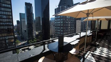 A rooftop bar with a checkered floor, a table with chairs, and a large umbrella.