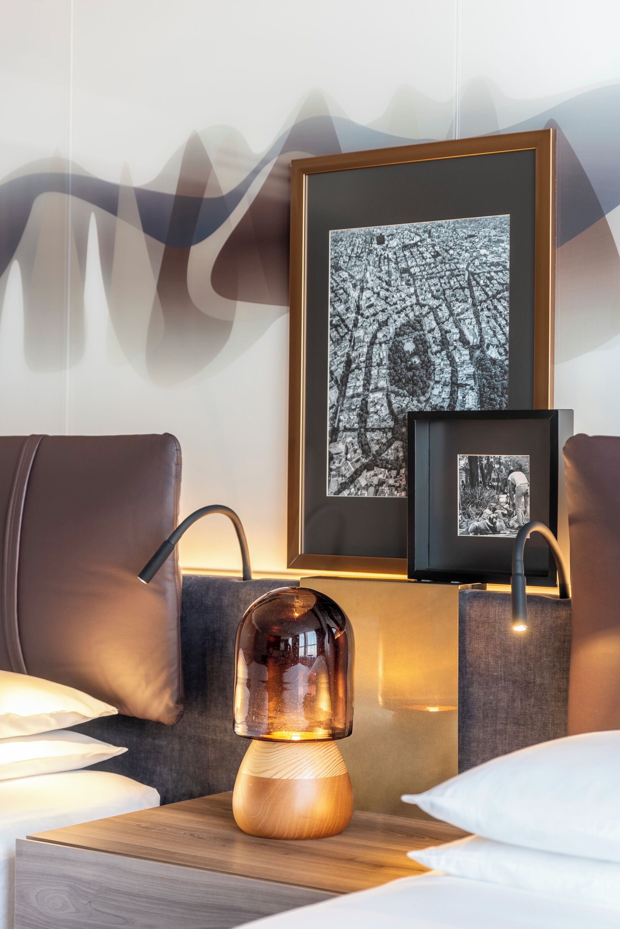 Une chambre d’hôtel moderne avec une table de chevet en bois, une lampe décorative, des photographies encadrées accrochées au mur et un fauteuil en cuir.