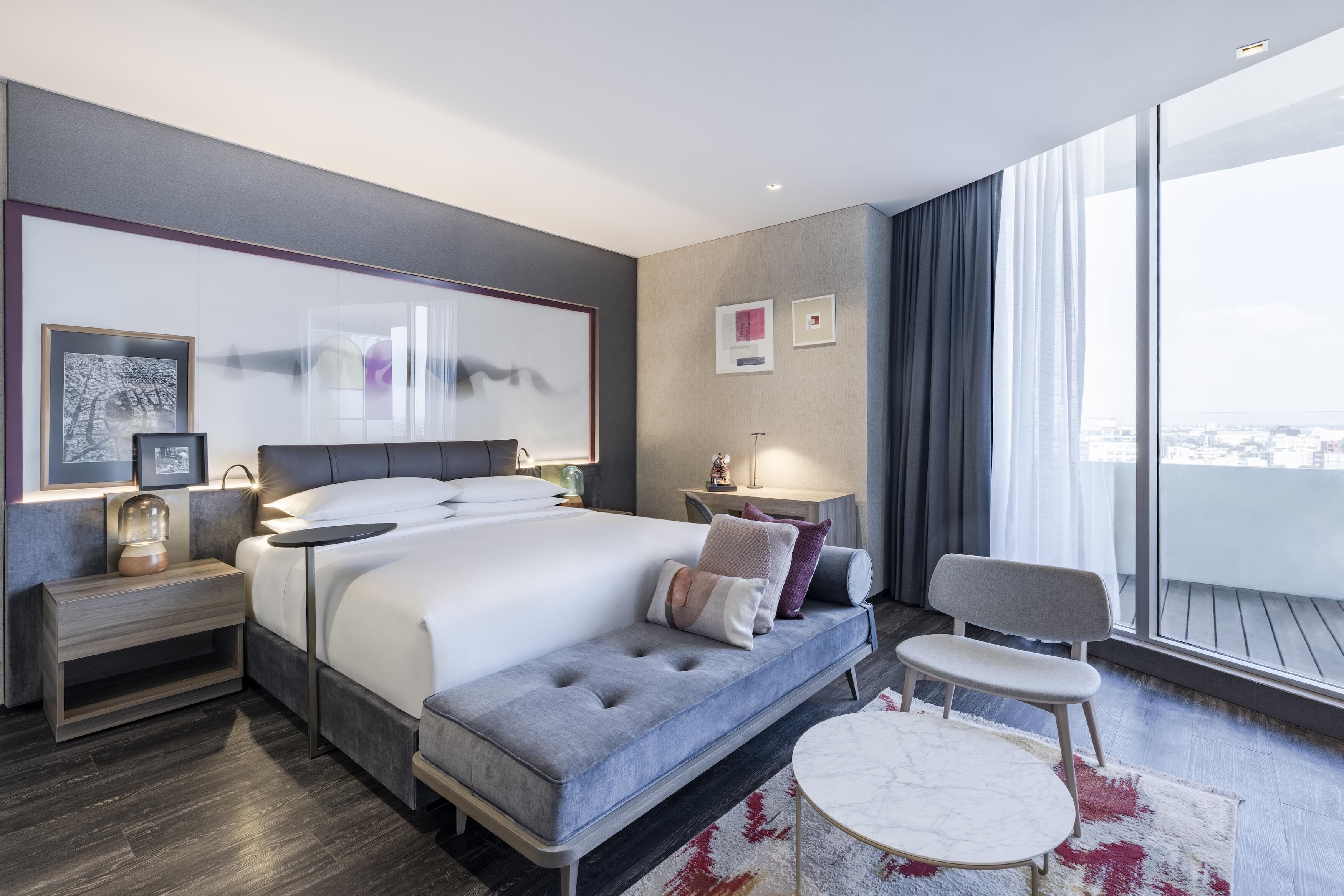 A modern hotel room with a large bed, bedside tables, a seating area, and a view through large windows.