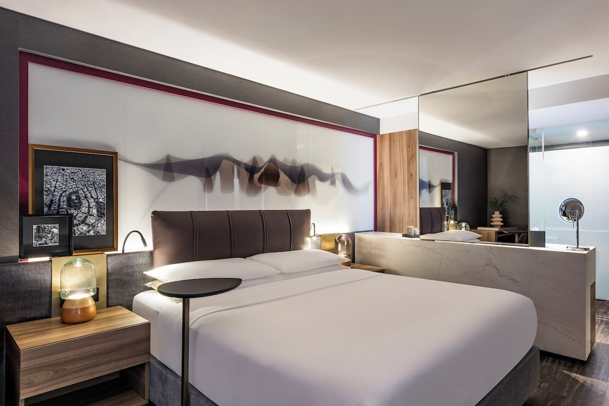 Une chambre d’hôtel moderne avec un grand lit, des tables de chevet, une petite table, une lampe fixée au mur et un grand miroir.