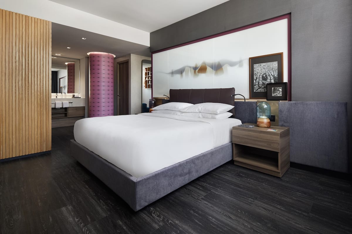 Une chambre d’hôtel moderne avec un grand lit, une table de chevet avec une lampe, un mur revêtu de panneaux en bois et un grand tableau abstrait.