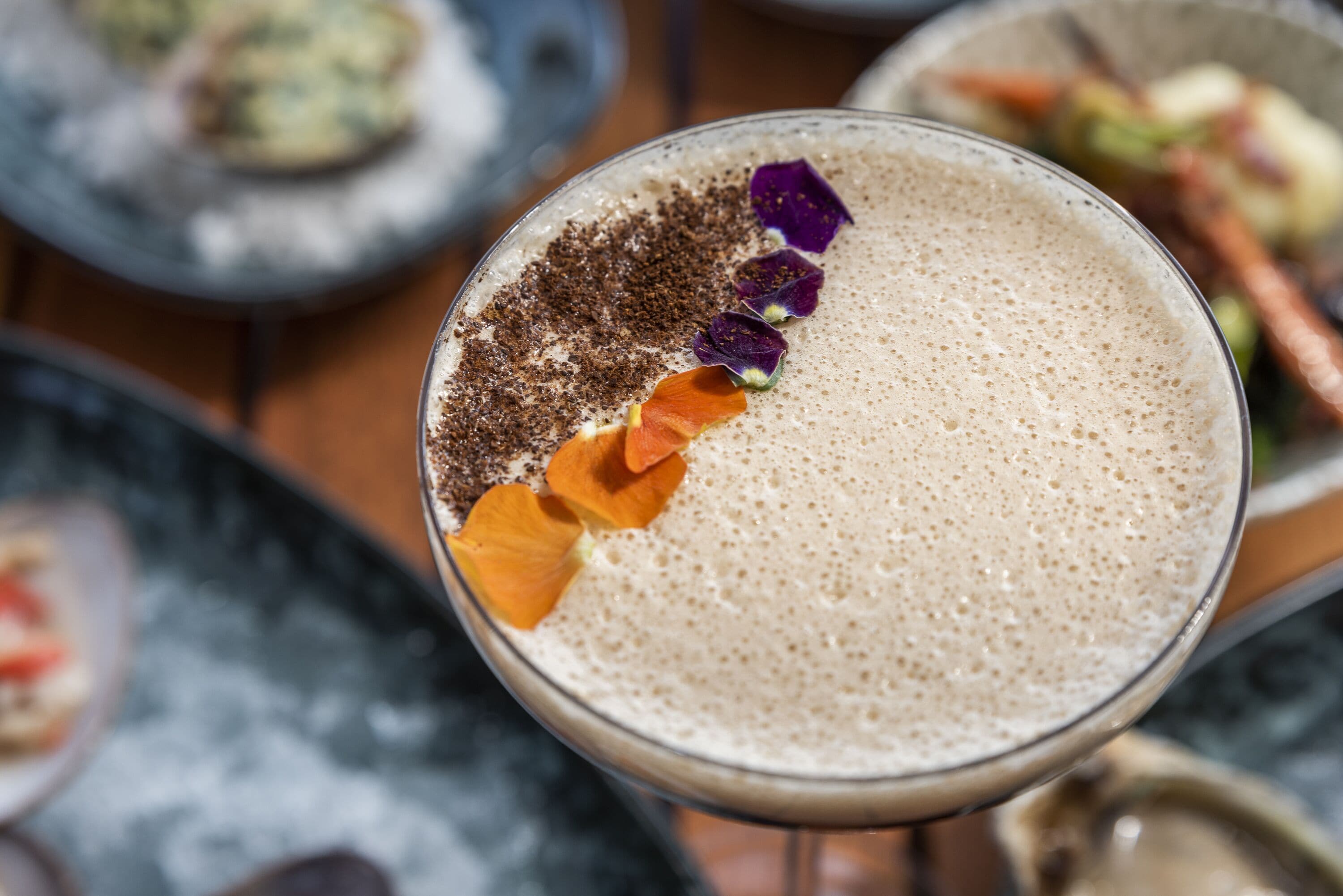 Gros plan d’une boisson mousseuse, décorée de poudre de chocolat, de zestes d’orange et de pétales de fleurs violettes.