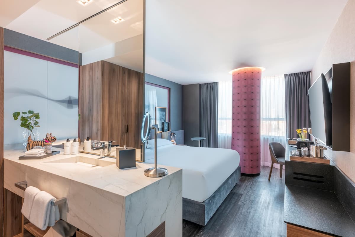 Une chambre d’hôtel moderne dotée d’un grand lit, d’une salle de bain avec un meuble-lavabo en marbre, d’un grand miroir et d’un mur d  ’accent de couleur rose.