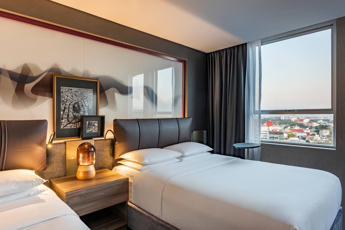 Une chambre d’hôtel avec un lit, des tables de chevet, une lampe, des œuvres d’art encadrées et une vue sur la ville.