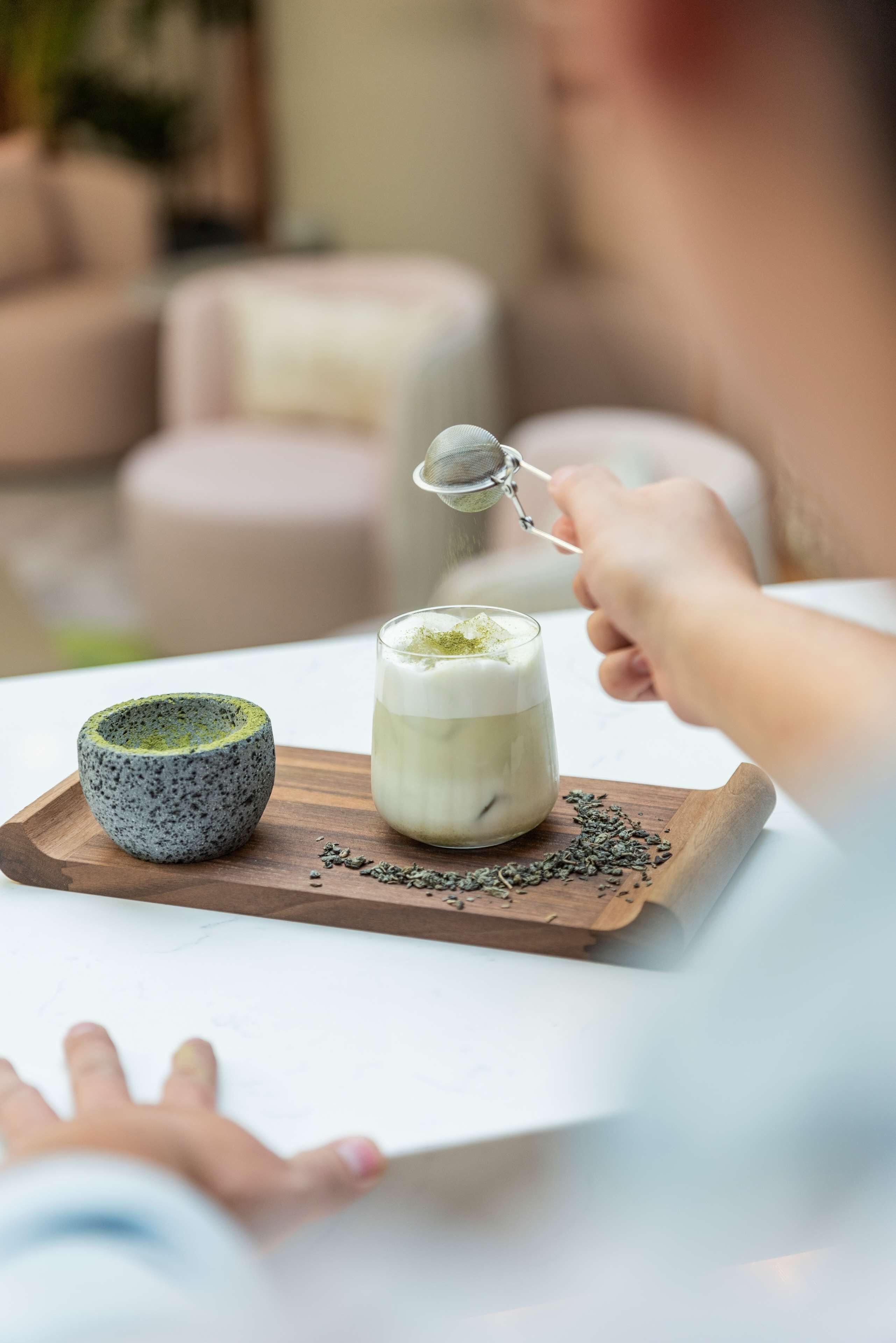 Una persona tiene un colino sopra un bicchiere di matcha su un vassoio di legno.
