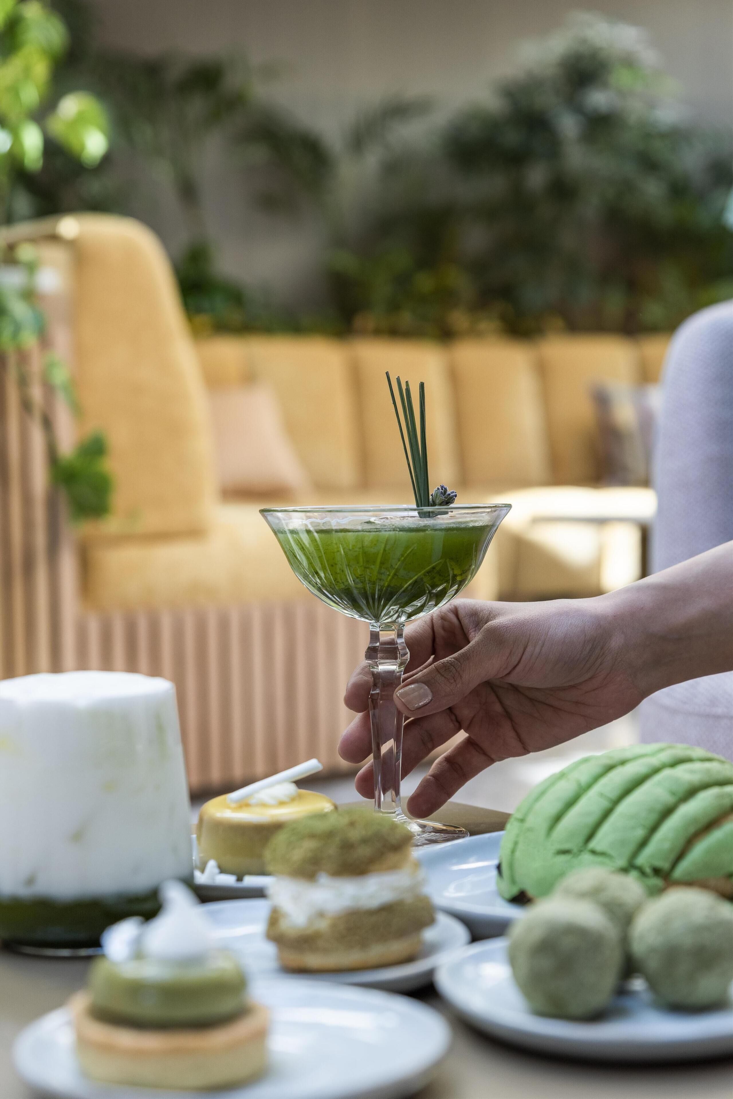 Una mano sosteniendo un vaso verde con cóctel y una pajita, rodeado de diversos postres sobre una mesa.