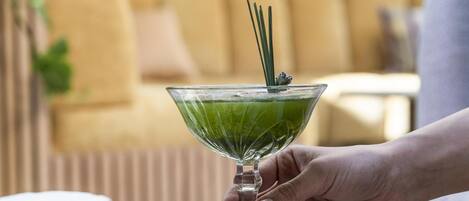 Una mano sosteniendo un vaso verde con cóctel y una pajita, rodeado de diversos postres sobre una mesa.