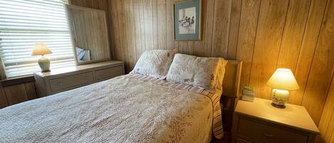 Un dormitorio con una pared revestida de madera, una cama con colcha estampada, una mesita de noche con lámpara y una ventana con persianas.