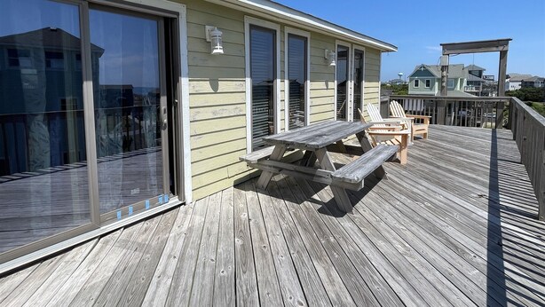 Une terrasse en bois avec une table de pique-nique et des chaises, une porte vitrée et un bâtiment doté de volets.