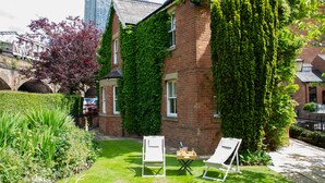 A brick house with ivy covering the walls, a lawn with two white lawn chairs, and a clear view of a tall building in the background. - Lock Keepers Cottage - Detached House in the City (Manchester)