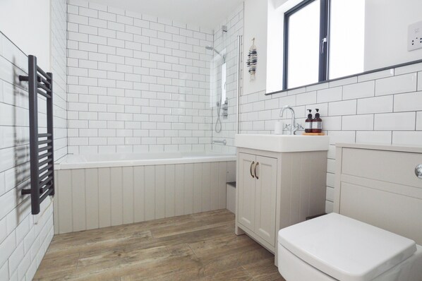Un baño con paredes de azulejos blancos, suelo de madera, una bañera blanca, un lavabo con espejo y un inodoro.