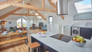 A spacious loft with a high ceiling, exposed wooden beams, a central island with a sink, a washing machine, and a dining area with a table and benches. - Birch House, situated just outside of the beautiful lively village of St Agnes and only 1.5 miles fr (St Agnes)
