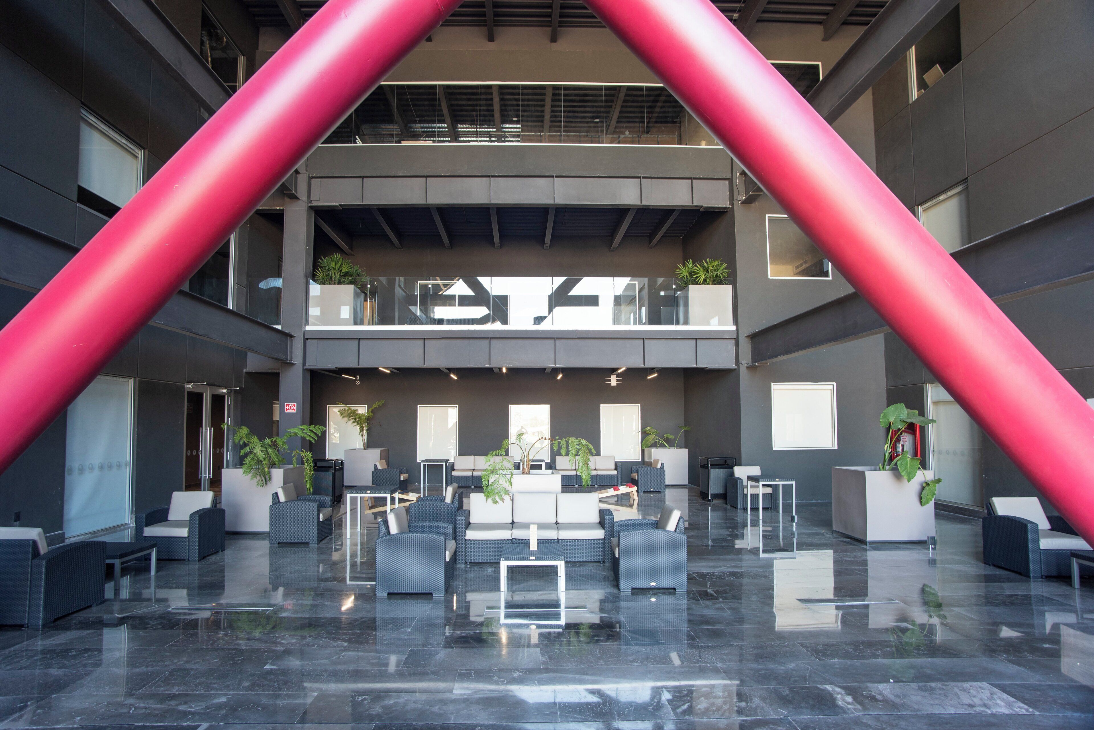 A spacious lobby with a modern design, featuring a central seating area, multiple potted plants, and a prominent red structural element.
