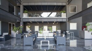 Un vestíbulo amplio con diseño moderno, que cuenta con una zona de estar central con sofás y sillones, un techo alto con vigas a la vista y plantas en macetas.