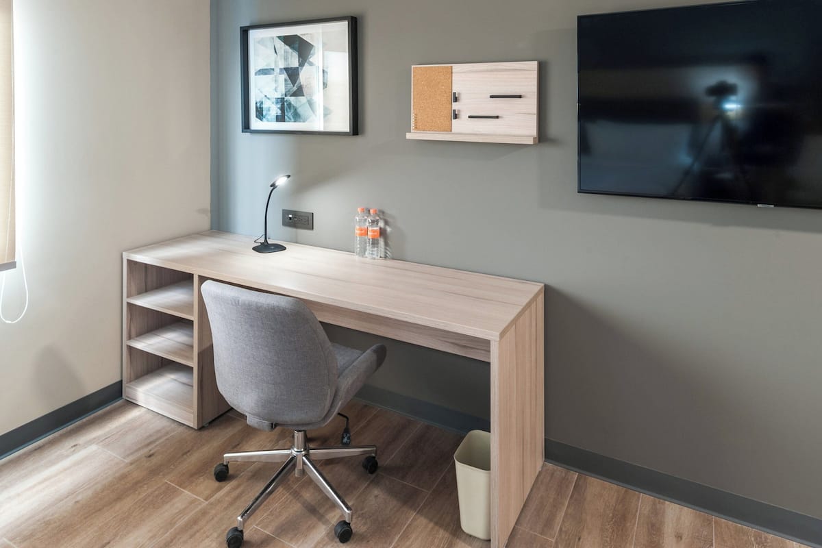 Ein moderner Arbeitsraum mit Schreibtisch aus Holz, einem grauen Bürostuhl, einem Wand-Fernseher und einem gerahmten Bild an der Wand.