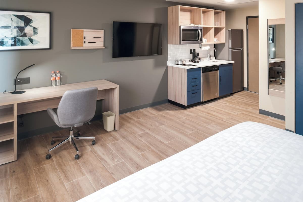 Ein modernes Hotelzimmer mit Schreibtisch aus Holz, einem grauen Bürostuhl, einem Bett, einer Küchenzeile mit Mikrowelle und einem Fernseher an der Wand.