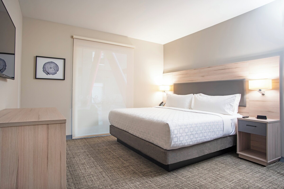 Quarto de hotel com cama, mesas de cabeceira, escrivaninha e uma janela grande com persianas.