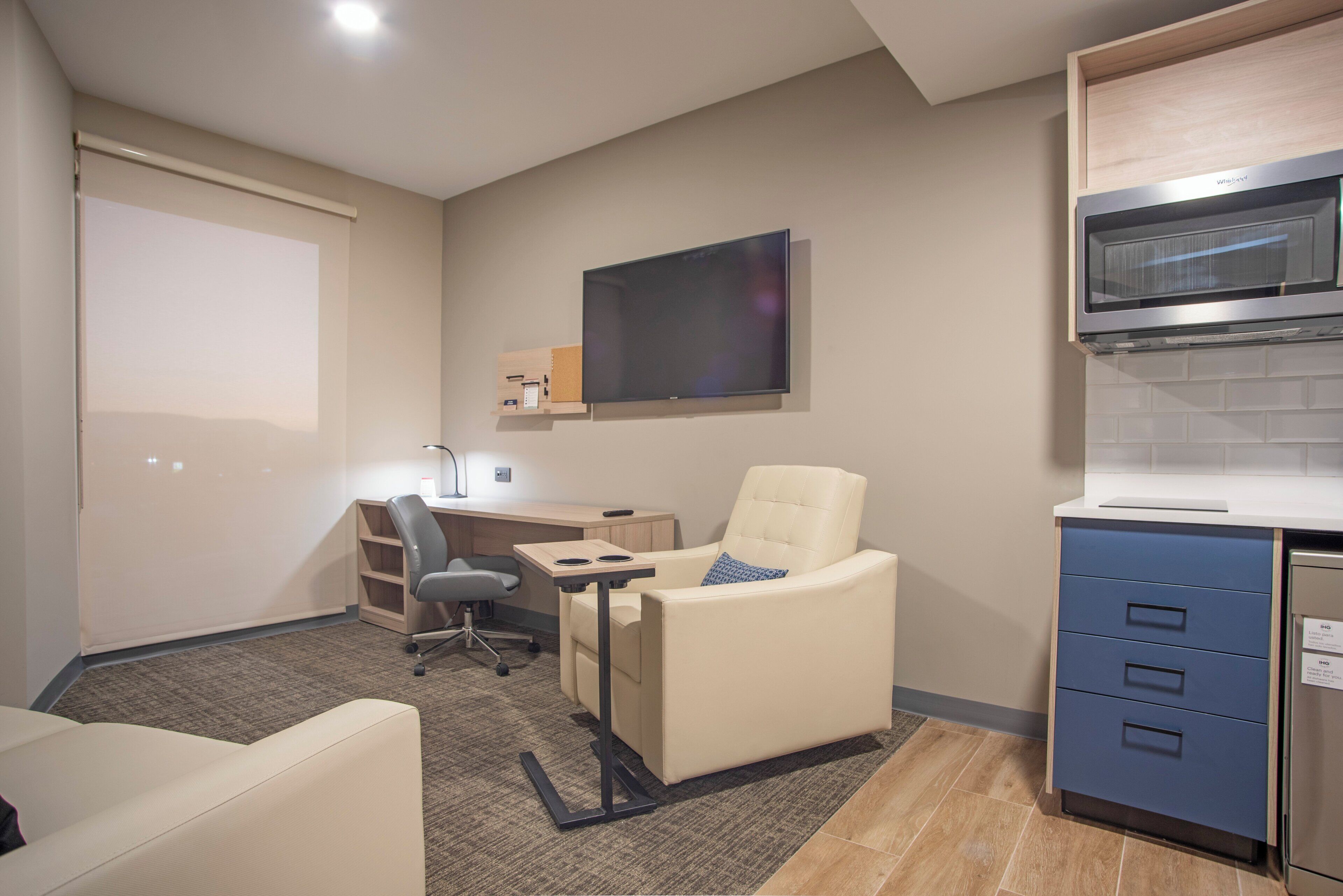 A modern room with a desk, a comfortable chair, a sofa, a flat-screen TV mounted on the wall, and a built-in kitchen area with a microwave.