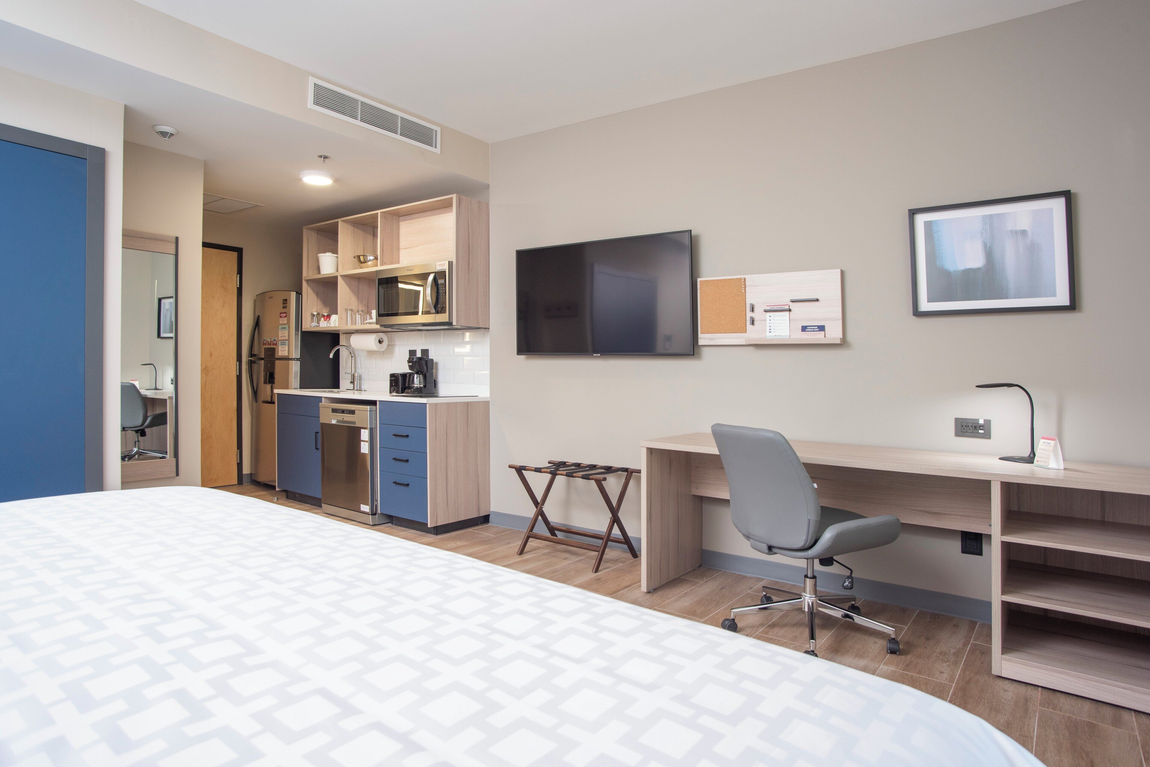 Quarto de hotel com cama, cozinha compacta, escrivaninha, cadeira e televisão.