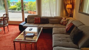 Un salón con sofá, una mesa de centro de vidrio y vistas a un jardín y montañas a través de amplios ventanales.