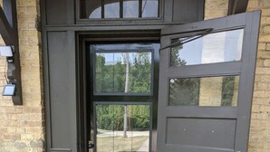 A dark brown door with a glass panel, a doormat with "Be our guest" text, and a brick wall. - Restored school house turned creative flex space (Perham)