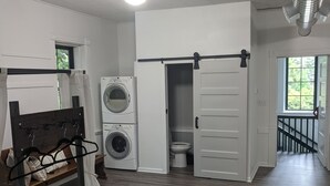 A hotel room with a bed, a washing machine, a toilet, and a sliding door leading to a balcony. - Restored school house turned creative flex space (Perham)