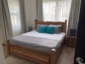 A neatly made bed with a wooden frame, a grey bedspread, and two teal pillows. There is a small bedside table with a lamp and a television.