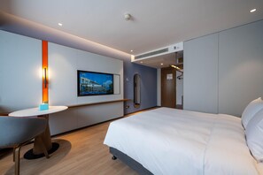 A modern hotel room with a bed, a desk with a chair, a television, and a wall-mounted artwork.