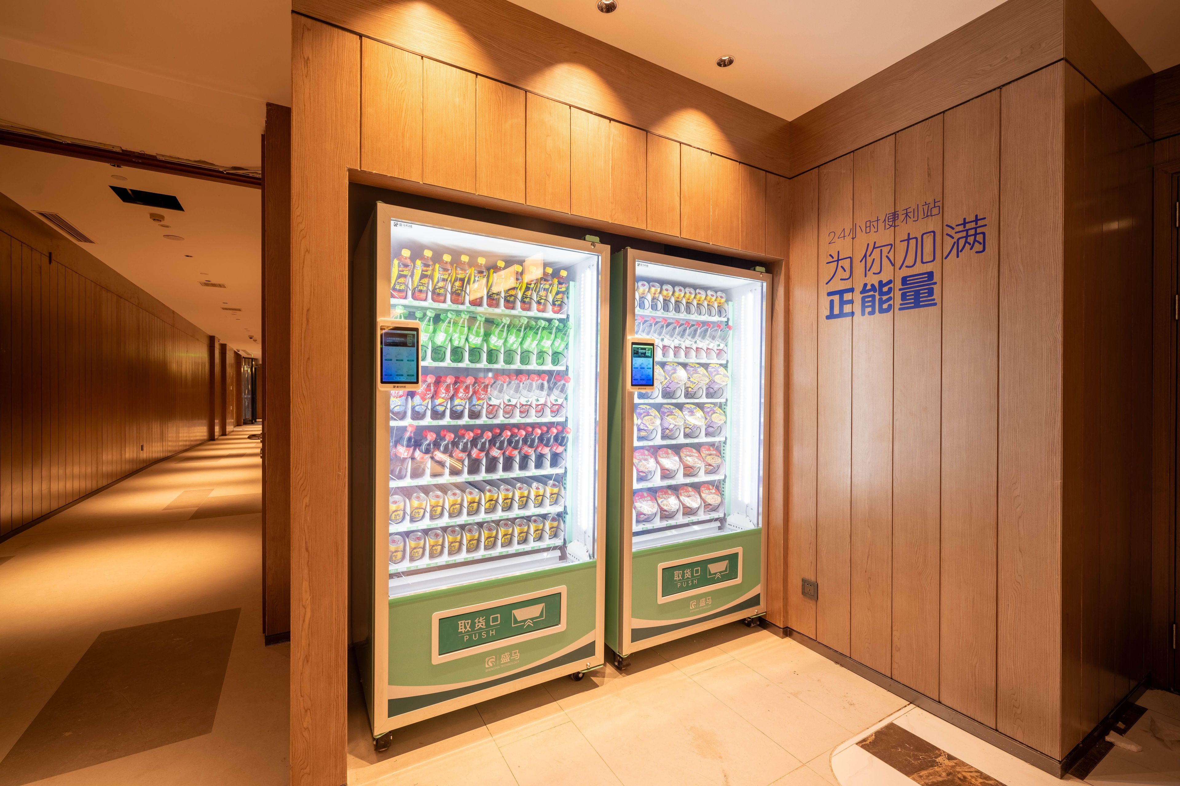 Um corredor com duas máquinas de venda automática repletas de bebidas e lanches, painéis de madeira nas paredes e uma placa com caracteres chineses.
