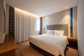 Una habitación de hotel moderna con una cama grande, una mesita de noche de madera, un escritorio integrado y un ventanal con cortinas.