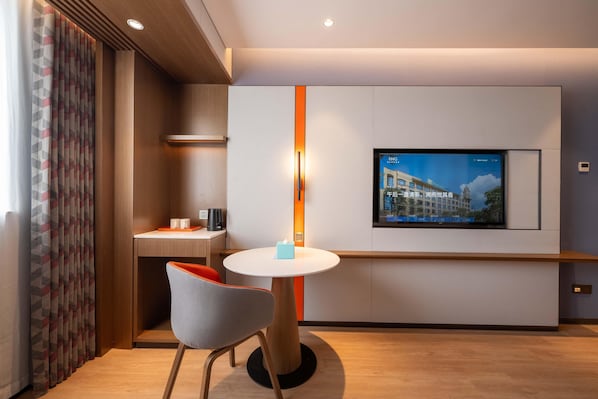 A modern hotel room with a flat-screen TV mounted on the wall, a small round table with a chair, and a desk with a chair.