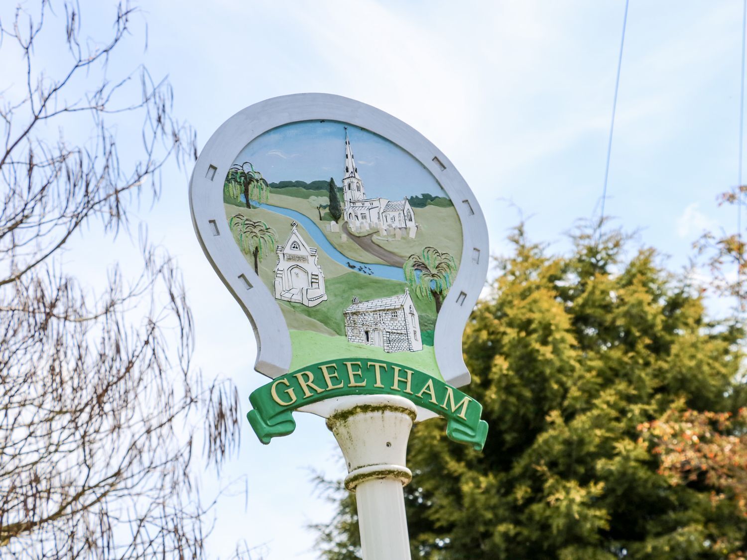 A signpost with a painted depiction of a village scene, including a church and houses, and the name 'GREETHAM' at the bottom.