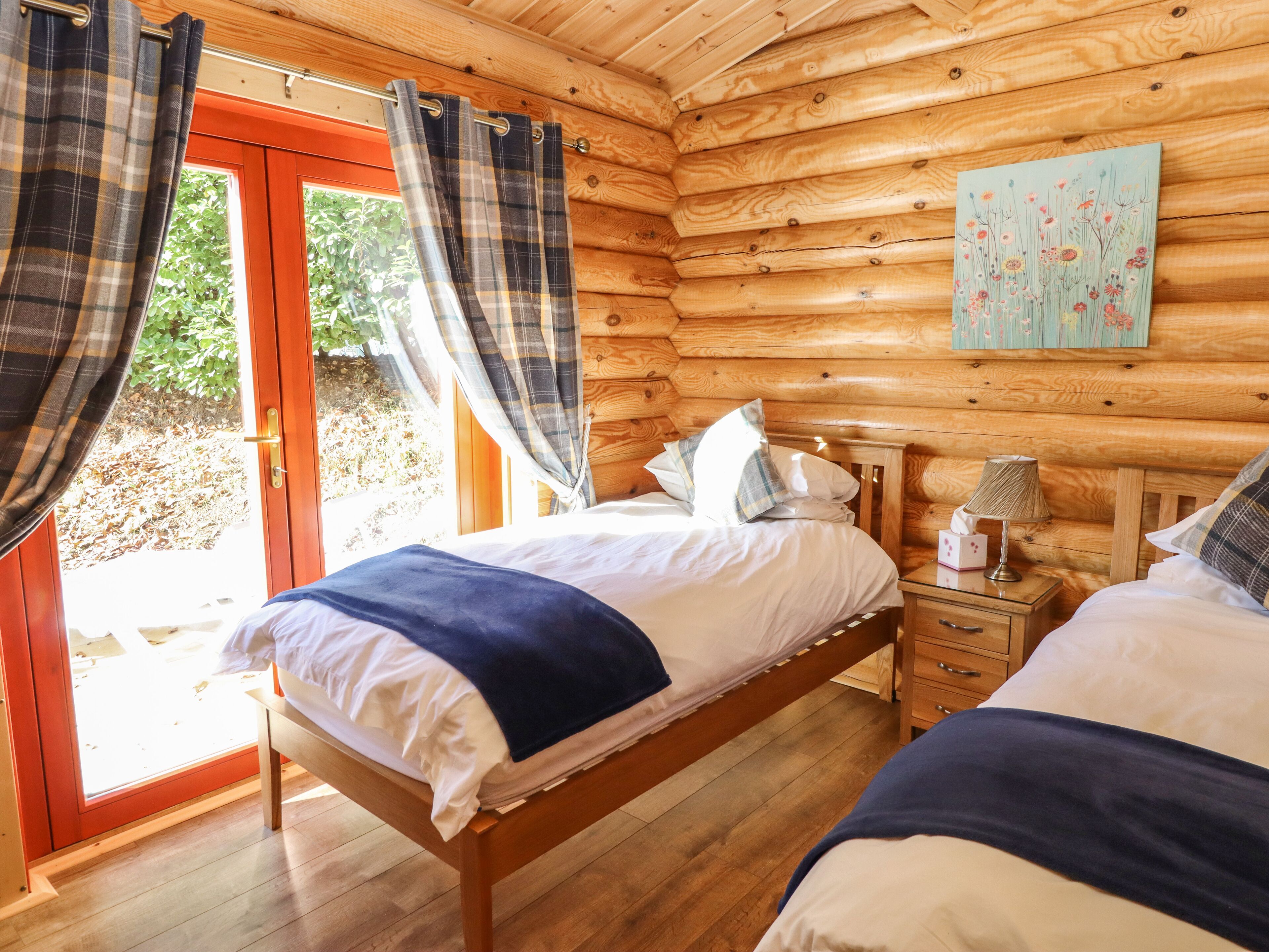 A wooden cabin bedroom with two beds, a wooden headboard, a nightstand with a lamp, and a painting on the wall.