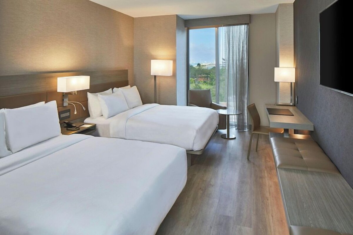 A hotel room with two beds, a desk, a chair, and a large window with curtains.