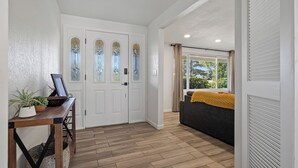 Ein Flur mit Holzboden, einer weißen Tür mit geätzten Glaseinsätzen, einem grauen Sofa mit gelber Decke und einem kleinen Tisch mit Pflanze und Bilderrahmen.