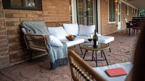 Eine Ziegelhausfassade mit überdachter Terrasse, auf der ein Korbstuhl, eine weiße Couch, ein Glastisch mit Obstschale und Wein sowie ein rotes Notizbuch zu sehen sind.