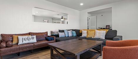 A modern living room with a brown leather sofa, a wooden coffee table, and a red armchair. There is a large mirror on the wall and a ceiling fan.
