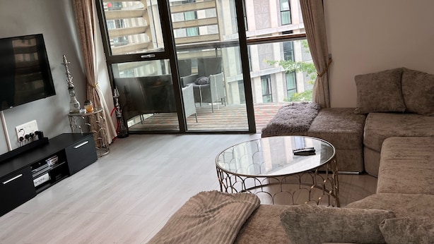 A modern living room with a sectional sofa, a glass coffee table, and a flat-screen TV on a black entertainment unit. - Stratford city apartment (London)