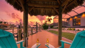 Um deck de madeira com cadeiras cor turquesa e duas garrafas de Corona, que dá acesso a um edifício com telhado de palha e janelas amplas.