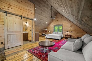 A cozy attic bedroom with a large bed, a sofa, a small table, and a window with a view of greenery.