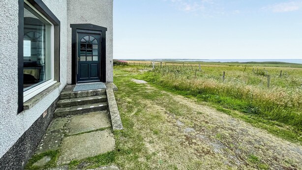 A house with a black door and windows, a stone staircase leading to the entrance, and a grassy field with a fence in the background. - The view rivals anywhere, it’s amazing. (Reiss, near Wick)
