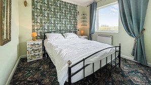 A bedroom with a bed, a nightstand, a lamp, a radiator, and a window with curtains. - The view rivals anywhere, it’s amazing. (Reiss, near Wick)