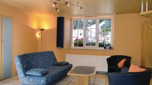 A cozy living room with a blue sofa, a small wooden table, and a black armchair. A window with a view of greenery and a radiator below it. - Awesome Home in Hachiville With Wifi and 1 Bedrooms (Wincrange)